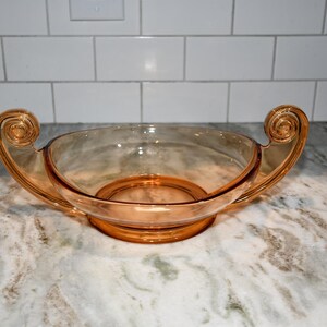 May include: A translucent amber-colored glass bowl featuring a wide, shallow basin and two prominent, upward-curving handles on opposite sides. Each handle terminates in a decorative spiral scroll design, and the bowl rests on a circular base.