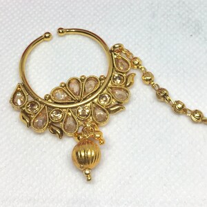 May include: A gold nose ring with a chain and a decorative design featuring clear stones. The ring is attached to a chain that hangs down from the ring.