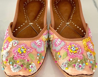 May include: A pair of pink embroidered flat shoes with colorful floral designs. The shoes have a brown leather insole and a pink satin upper.