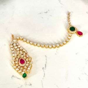 Może przedstawiać: Złoty łańcuszek do nosa z wisiorem w kształcie łzy ozdobionym przezroczystymi i różowymi cyrkoniami. Łańcuszek jest ozdobiony przezroczystymi cyrkoniami i ma dwa zielone akcenty w kształcie łzy.