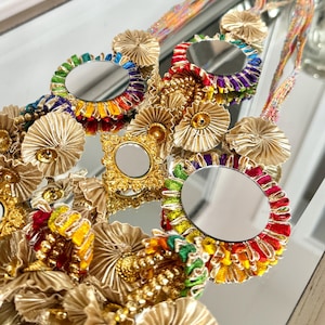 May include: A decorative necklace featuring small round and square mirrors with gold frames. The mirrors are surrounded by colorful, ruffled trim in rainbow hues. Gold fabric flowers and ribbons are also incorporated into the design.