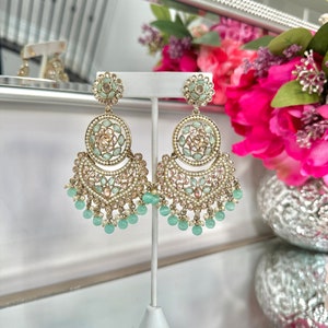 May include: A pair of gold tone dangle earrings with a multi-tiered design. The earrings feature a large round center piece with green and pink stones. The bottom tier features a row of green beads.