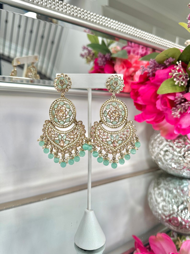 May include: A pair of gold tone dangle earrings with a multi-tiered design. The earrings feature a large round center piece with green and pink stones. The bottom tier features a row of green beads.