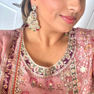 May include: A woman wearing a pink and purple patterned dress with gold embroidery and a pair of large, ornate gold earrings with colorful stones.