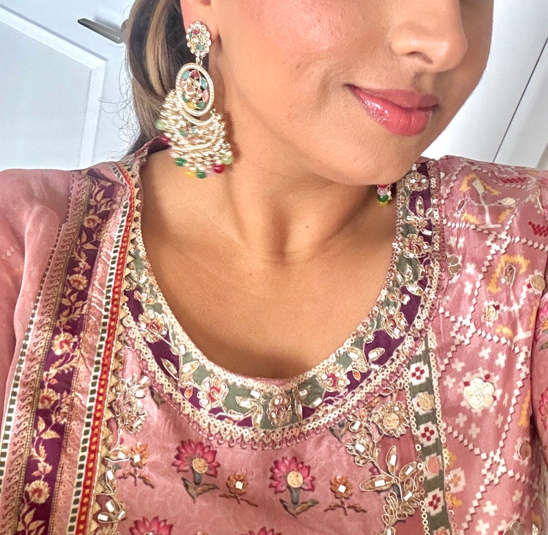 May include: A woman wearing a pink and purple patterned dress with gold embroidery and a pair of large, ornate gold earrings with colorful stones.