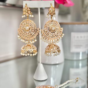 May include: Gold-toned chandelier earrings with intricate detailing. Each earring features a large, circular design with a central pearl, surrounded by smaller pearls and ornate embellishments. The earrings have a bell-shaped bottom, also adorned with pearls.