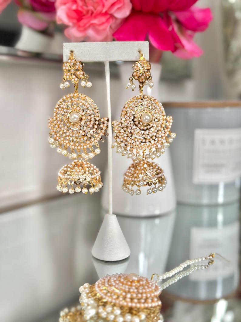 May include: Gold-toned chandelier earrings with intricate detailing. Each earring features a large, circular design with a central pearl, surrounded by smaller pearls and ornate embellishments. The earrings have a bell-shaped bottom, also adorned with pearls.