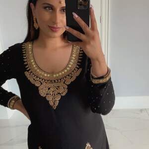 Op de afbeelding: Een vrouw draagt een zwarte outfit met gouden borduurwerk. Ze draagt een gouden hoofdtooi en oorbellen. Ze houdt een zwarte smartphone in haar hand.
