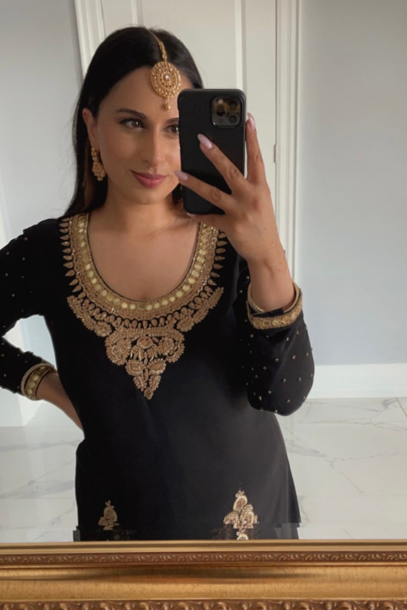 Op de afbeelding: Een vrouw draagt een zwarte outfit met gouden borduurwerk. Ze draagt een gouden hoofdtooi en oorbellen. Ze houdt een zwarte smartphone in haar hand.