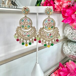 May include: A pair of gold earrings with multi-colored stones and pearls. The earrings are hanging from a stand and have a crescent moon shape with a circle in the center.