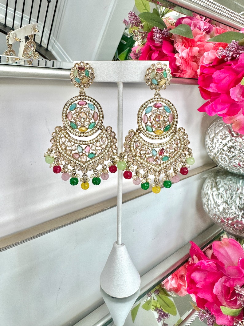 May include: A pair of gold earrings with multi-colored stones and pearls. The earrings are hanging from a stand and have a crescent moon shape with a circle in the center.