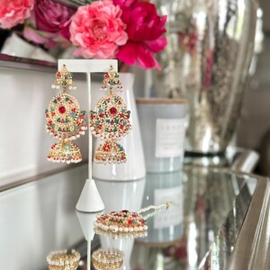 May include: Gold-toned, ornate earrings with red, green, and white gemstones. The earrings are displayed on a white stand, alongside a matching pendant and ring. The jewelry reflects light, creating a sparkling effect. A floral arrangement is in the background.