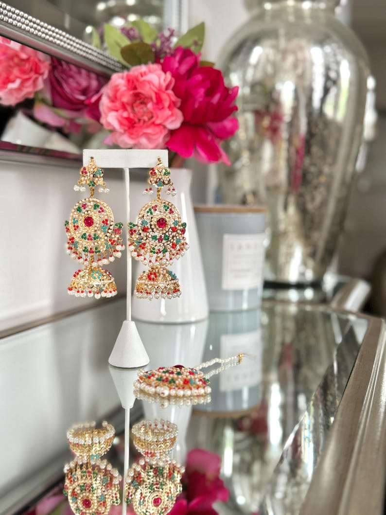 May include: Gold-toned, ornate earrings with red, green, and white gemstones. The earrings are displayed on a white stand, alongside a matching pendant and ring. The jewelry reflects light, creating a sparkling effect. A floral arrangement is in the background.
