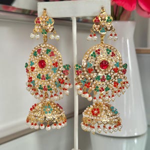 May include: Gold-toned chandelier earrings with intricate detailing. Each earring features a floral top, a circular center with a red stone, and a bell-shaped bottom. Embellished with colorful accents and dangling pearls, these earrings are displayed on a white stand.