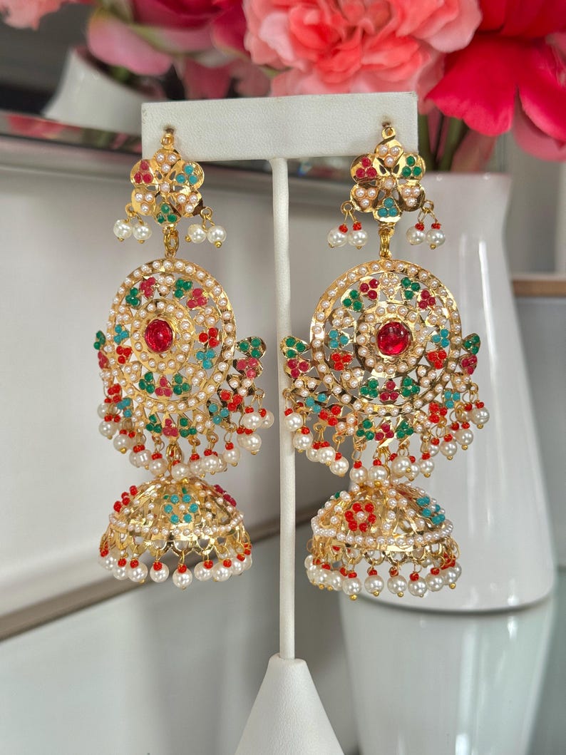 May include: Gold-toned chandelier earrings with intricate detailing. Each earring features a floral top, a circular center with a red stone, and a bell-shaped bottom. Embellished with colorful accents and dangling pearls, these earrings are displayed on a white stand.