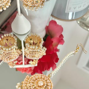 May include: Gold-toned jewelry set featuring earrings and a headpiece, adorned with small pearls. The earrings have a jhumka style with a pearl-studded dome. The headpiece has a central pearl and a pearl strand. The set is displayed on a reflective surface.