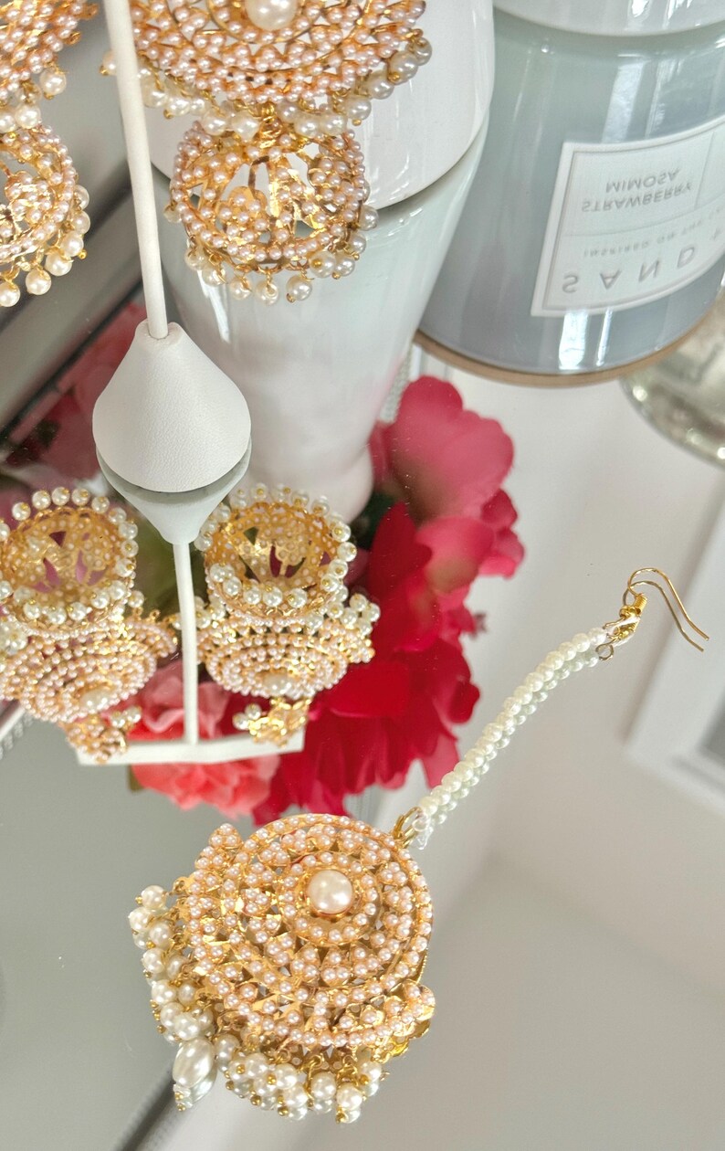 May include: Gold-toned jewelry set featuring earrings and a headpiece, adorned with small pearls. The earrings have a jhumka style with a pearl-studded dome. The headpiece has a central pearl and a pearl strand. The set is displayed on a reflective surface.