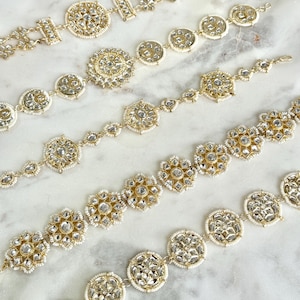 May include: Several gold-toned, ornate bracelets with intricate designs. Each bracelet features a series of circular and floral-shaped embellishments, adorned with clear, sparkling stones and small white beads. The bracelets are displayed on a marble surface.