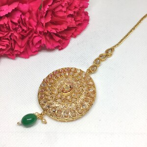 Op de afbeelding: Gouden maang tikka met een ronde, sierlijke hanger bezet met heldere en amberkleurige strass steentjes. De hanger is bevestigd aan een delicate ketting met een groene kraal aan het einde.