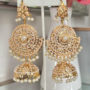 May include: Gold-toned earrings with intricate detailing. Each earring features a floral top, a circular design with small pearls, and a bell-shaped bottom also adorned with pearls. The earrings are displayed on a white stand.