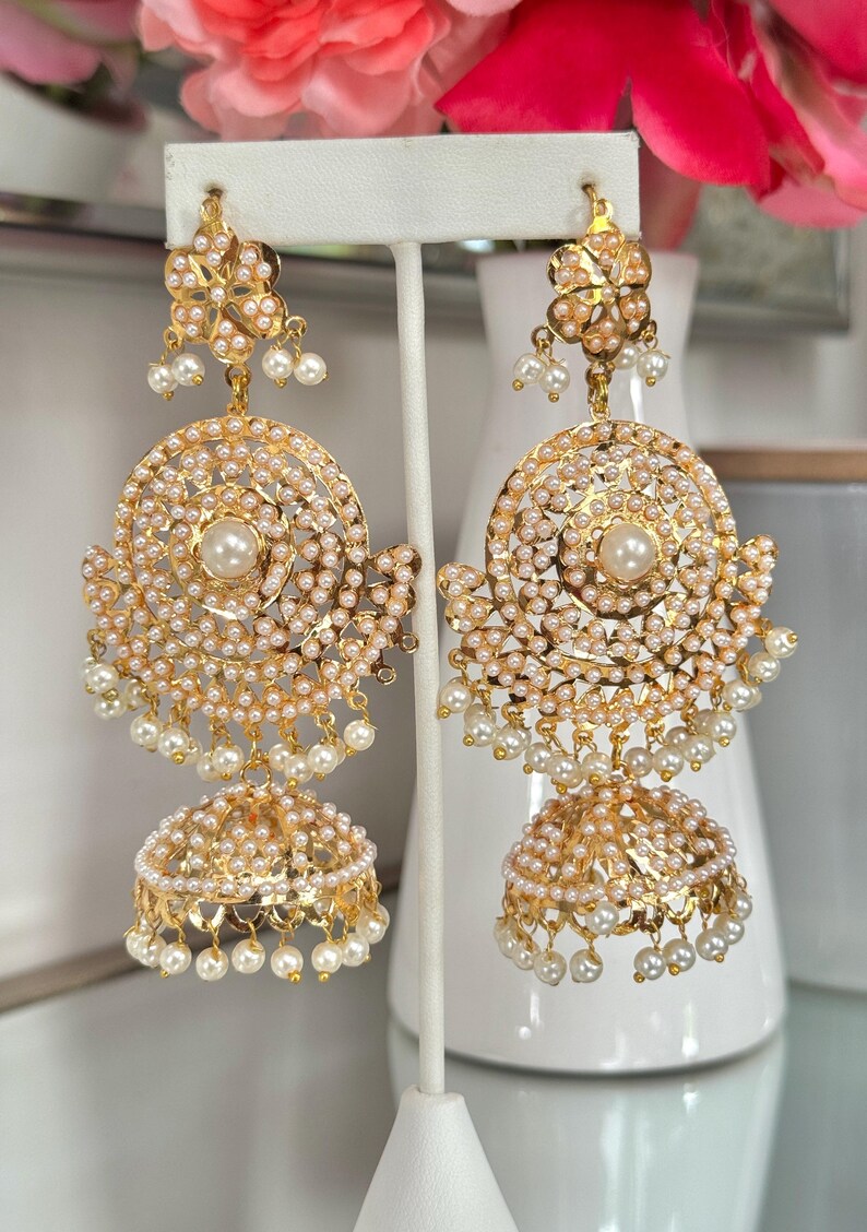 May include: Gold-toned earrings with intricate detailing. Each earring features a floral top, a circular design with small pearls, and a bell-shaped bottom also adorned with pearls. The earrings are displayed on a white stand.