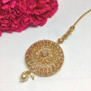 Op de afbeelding: Een gouden maang tikka met een ronde, sierlijke hanger met heldere en amberkleurige stenen. De hanger is bevestigd aan een delicate ketting met een parel.
