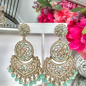 May include: A pair of gold tone earrings with a large, round, multi-layered design. The earrings are adorned with light green and clear stones and have a fringe of light green beads.