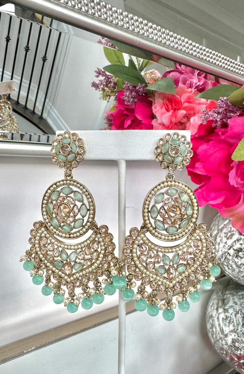 May include: A pair of gold tone earrings with a large, round, multi-layered design. The earrings are adorned with light green and clear stones and have a fringe of light green beads.
