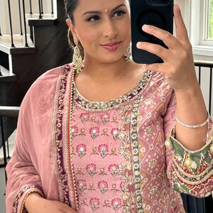 May include: A woman wearing a pink and green embroidered dress with gold embellishments. She is holding a black smartphone in her right hand and has a silver bangle on her left wrist.