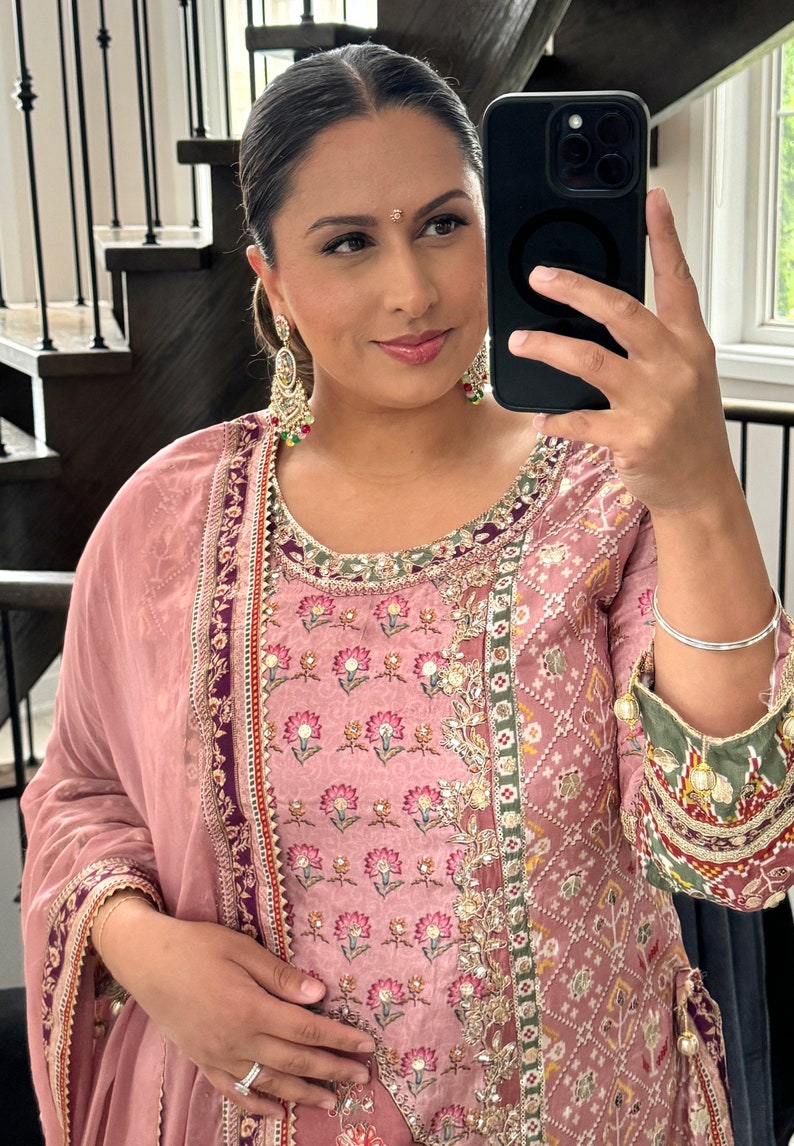 May include: A woman wearing a pink and green embroidered dress with gold embellishments. She is holding a black smartphone in her right hand and has a silver bangle on her left wrist.