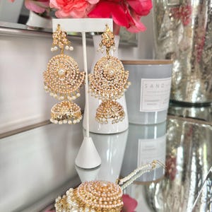 May include: Gold-toned, ornate earrings with pearl accents. The earrings feature a layered design with a central pearl and dangling pearls. The image also shows a matching bracelet, a white earring stand, and a vase of pink flowers.