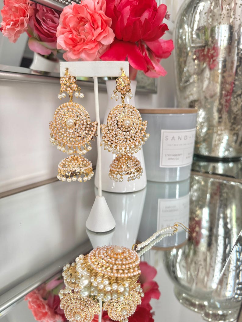 May include: Gold-toned, ornate earrings with pearl accents. The earrings feature a layered design with a central pearl and dangling pearls. The image also shows a matching bracelet, a white earring stand, and a vase of pink flowers.