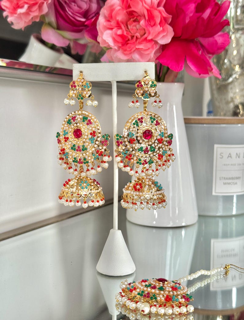 May include: Gold-toned chandelier earrings with red, green, and turquoise accents, and dangling white pearls. The earrings feature a floral design and are displayed on a white stand. A matching pendant is also visible.
