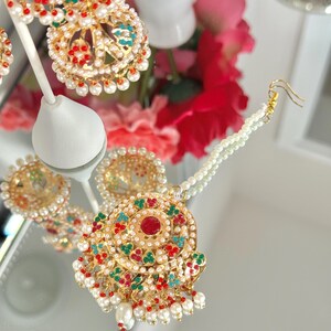May include: Close-up of gold-toned jewelry with red, green, and turquoise accents. The pieces are adorned with small white pearls. The jewelry includes earrings and a pendant, all with intricate designs and a vintage aesthetic.