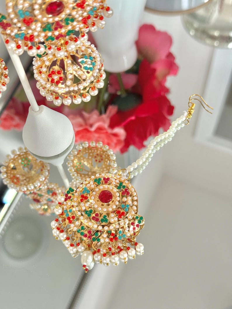 May include: Close-up of gold-toned jewelry with red, green, and turquoise accents. The pieces are adorned with small white pearls. The jewelry includes earrings and a pendant, all with intricate designs and a vintage aesthetic.
