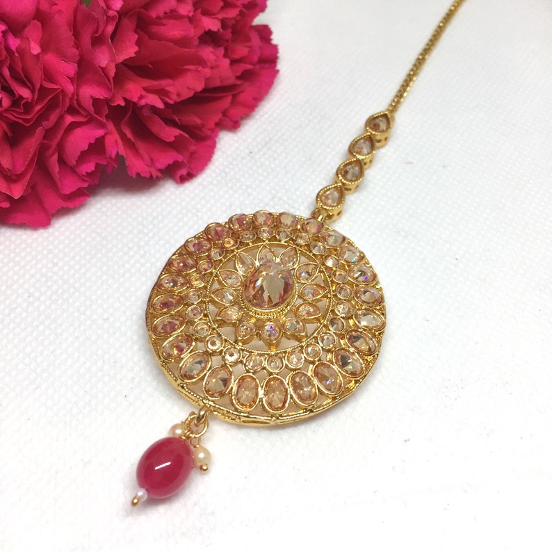 Op de afbeelding: Een gouden maang tikka met een ronde, sierlijke hanger met heldere en amberkleurige stenen. De hanger is bevestigd aan een ketting met heldere stenen en eindigt met een rode kraal.