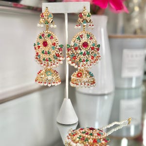 May include: Gold-toned chandelier earrings with red, green, and white accents. The earrings feature a large, circular design with a central red stone and dangling pearl-like beads. A matching headpiece is also visible.