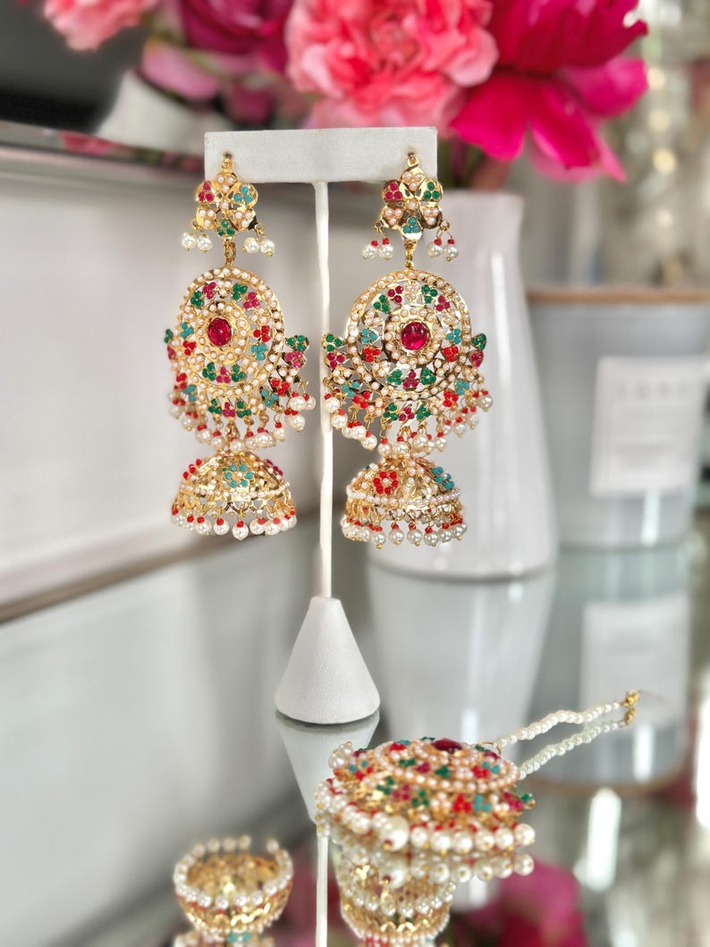 May include: Gold-toned chandelier earrings with red, green, and white accents. The earrings feature a large, circular design with a central red stone and dangling pearl-like beads. A matching headpiece is also visible.