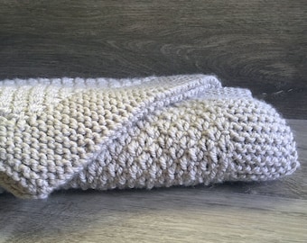 May include: A soft, grey knitted blanket folded on a wooden surface. The blanket is made of a textured yarn and has a subtle pattern.