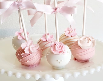 Puede incluir: Un soporte blanco para pasteles contiene varios cake pops. Los cake pops están decorados con glaseado rosa, flores y lazos. Algunos son blancos con detalles en rosa, otros rosas con detalles en blanco. Los cake pops están en palitos blancos.