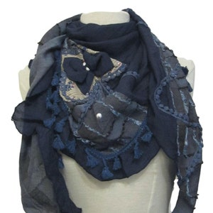 Puede incluir: Bufanda azul marino con diseño patchwork. La bufanda presenta varias texturas, incluyendo borlas, cuentas y tela estampada. La bufanda está drapeada sobre un maniquí, mostrando su forma y detalles.