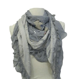 Könnte beinhalten: Grauer Schal mit einem mehrschichtigen Design, präsentiert auf einer Schaufensterpuppe. Der Schal hat ein Patchwork-Design mit floralen Akzenten und kleinen Perlenverzierungen. Die Kanten sind mit Quasten und Rüschen verziert, was einen strukturierten, femininen Look erzeugt.