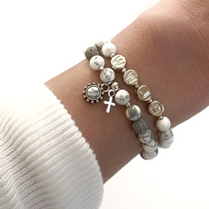 May include: Two bracelets with white and gray beads. One bracelet has a silver cross charm and a silver flower charm. The bracelets are on a person's wrist.