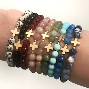 May include: A set of eight bracelets with different colored gemstone beads and gold cross charms. The bracelets are stacked on a person's wrist.