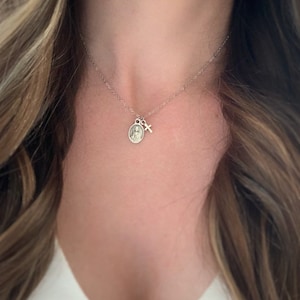 May include: A silver chain necklace with a small silver cross charm and a silver oval charm with a religious image.