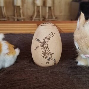 May include: A wooden egg-shaped ornament with a Celtic knot design of a rabbit. The ornament is sitting on a brown furry surface with two white and brown bunnies.