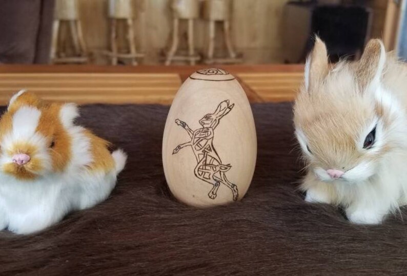 May include: A wooden egg-shaped ornament with a Celtic knot design of a rabbit. The ornament is sitting on a brown furry surface with two white and brown bunnies.