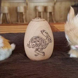May include: A wooden egg-shaped ornament with a Celtic knot design of a rabbit. The ornament is sitting on a brown fur surface with two white and brown fluffy bunnies.