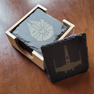 May include: Set of six black slate coasters with a wooden holder. The coasters feature laser-etched images of the Millennium Falcon and X-Wing starfighters from Star Wars.