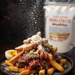 Puede incluir: Un plato de patatas fritas cargadas con carne, salsa, semillas de s&eacute;samo y cebolletas. Se espolvorea polvo blanco sobre las patatas fritas. Hay media rodaja de huevo duro a un lado. Una bolsa de "Sweet Korean Snow Cheese" est&aacute; en el fondo.